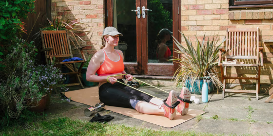 Woman sitting on a yoga may, exercising in her garden, using a resistance band