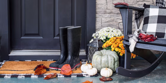 Traditional style front porch decorated for autumn with rain boots, heirloom gourds, white pumpkins, mums and rocking chair with buffalo plaid pillow and throw blanket giving an inviting atmosphere.
