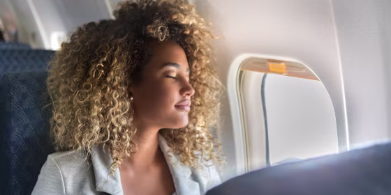 A young woman sits in the window seat of a commercial airliner and leans back with her eyes closed.