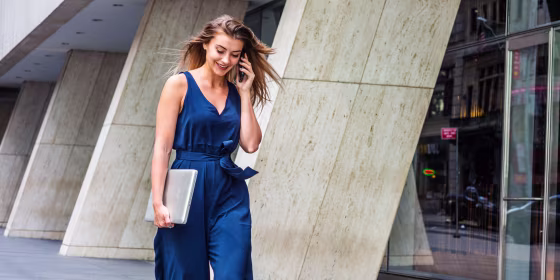 Young Eastern European American Woman talking on cell phone, traveling, working in New York City, wearing blue sleeveless jumpsuit, carrying laptop computer, walking on street outside office building