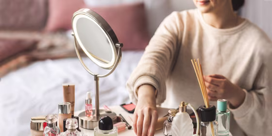Cosmetics are laid out on a round white table. Beautiful little glass bottles and tubes with shiny lids, and some perfume. Woman is in the process of doing her make up.