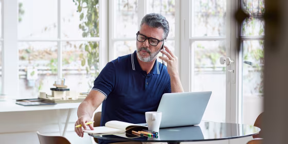 Mature businessman using mobile phone while writing notes at table in office