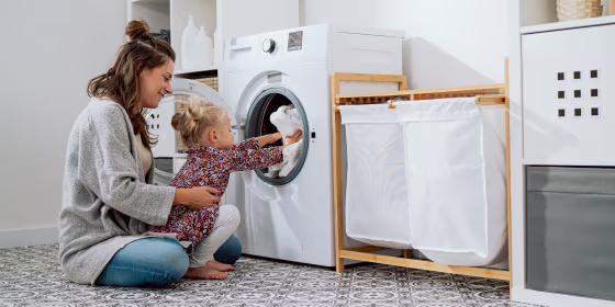 A caring mom with her little daughter sits in the bathroom, laundry at the washing machine, a sweet little girl sits on a woman's lap and puts towels in the open drum