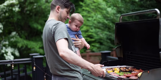 A man holding a baby, while grilling some meat and vegetables