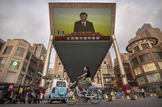 A woman wearing a face mask rides a bicycle past a large television screen at a shopping center displaying Chinese state television news coverage of