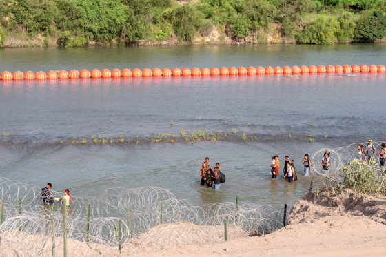 Migrants walk by a string of buoys placed on the water along the Rio Grande border with Mexico in Eagle Pass, Texas, on July 16, 2023. The buoy installation is part of an operation Texas is pursuing to secure its borders, but activists and some legislators say Governor Greg Abbott is exceeding his authority.