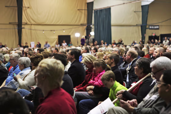 An auditorium is filled with people sitting in chairs, listening to an unseen speaker 