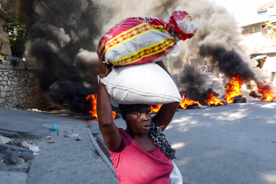 A woman carrying two bags of rice walks past burning tires