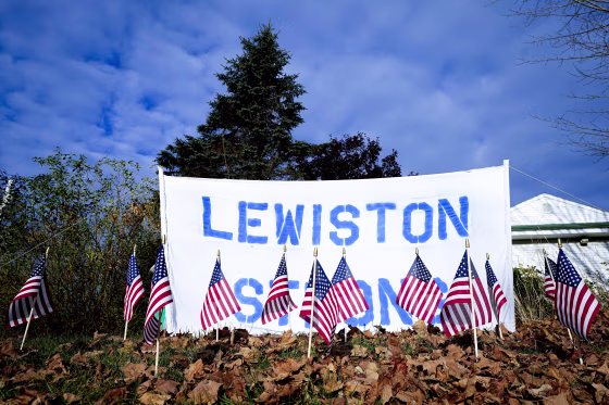 A white poster with blue text reads "Lewiston Strong" behind American flags posted in the grass lawn in front of it