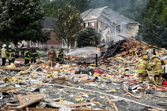 A damaged house and explosion debris outside, firefighters use a hose to put out the fire