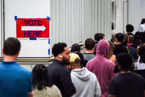 People line up to vote, a "Vote Here Sign" is taped to a wall nearby