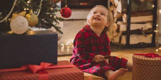A 1 year old childing in plaid pajamas sitting under a Christmas tree
