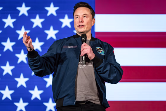 Elon Musk speaks on stage, with an American flag backdrop behind him