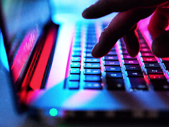Fingers typing on laptop keyboard at night, illuminated by red, blue, and pink lights