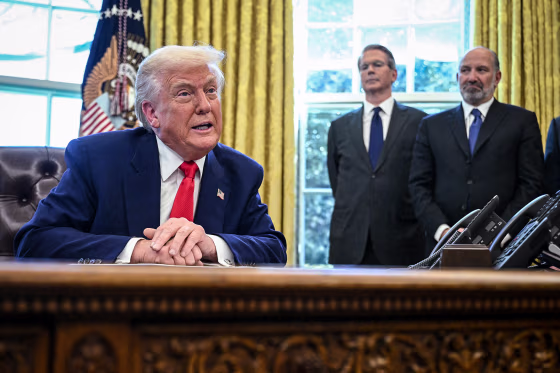 President Donald Trump, Treasury Secretary Scott Bessent, and Commerce Secretary Howard Lutnick inside of the Oval Office, Trump is seated at a desk, the other two are standing behind him
