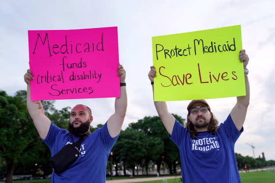 Two people hold signs outside, one the left it says "Medicaid funds critical disability services", and on the right it says "Protect Medicaid, Save lives"