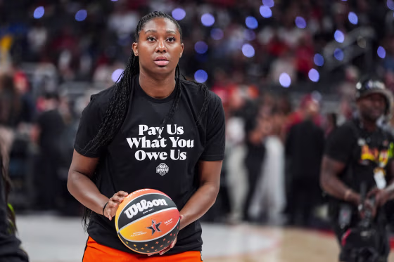 Aliyah Boston holds a basketball on the court, wearing a black shirt with white lettering that reads "Pay Us What You Owe Us"