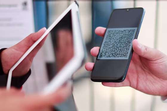 A person holding a tablet uses it to scan a QR code displayed on someone's cellphone