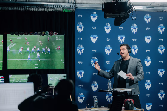 A person wearing headphones standing in front of a blue backdrop on set speaks near a television screen displaying a football game