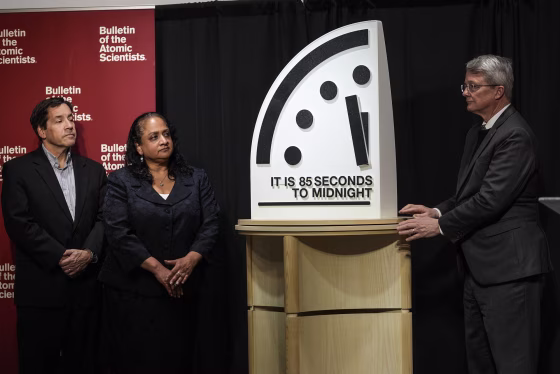 Three people stand to either side of an oversized clock sculpture with text reading "IT IS 85 SECONDS TO MIDNIGHT".