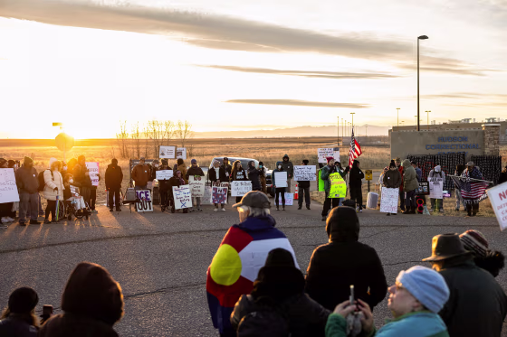 The sun rises in the distance and throws warm light across a crowd of protesters holding signs and bundled against the cold.