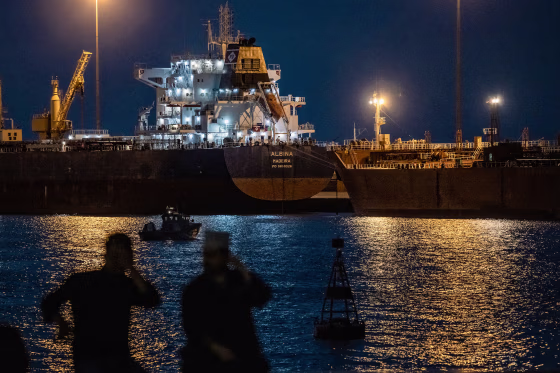 Two men are silhouetted against lights from large tankers floating across the water at night.