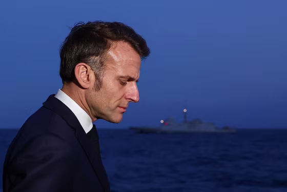 Emmanuel Macron appears in profile view in front of a battleship floating in the sea at dusk.