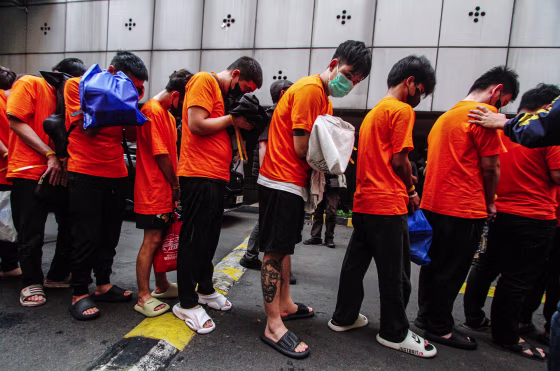 People wearing orange t-shirts with orange zip ties binding their wrists are walked by police in a line
