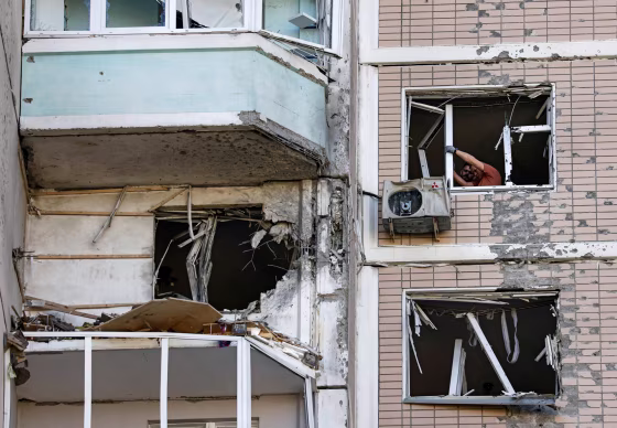 Residents clear broken windows in a damaged apartment building, as seen from the outside of the building