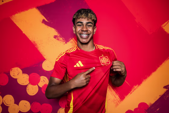 Lamine Yamal smiles and points to the icon for Spain on his soccer jersey in front of a red colored background