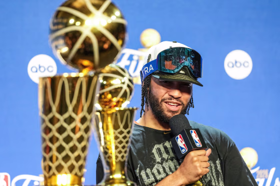 Jalen Brunson speaks white seated during a press conference, two NBA Finals winner trophies are placed in front of him