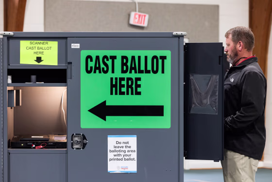 James Couch votes at a voting booth inside of a room, a large green sign that reads "cast ballot here" in black text is on the side of the voting booth