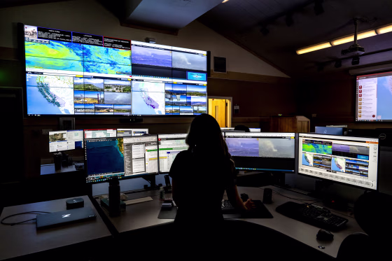 A Cal Fire worker monitors computers showing wildfire camera systems in a dark room.