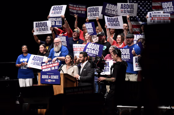 Abdul El-Sayed and Alexandria Ocasio-Cortez on stage during a campaign rally, surrounded by people holding ABDUL FOR SENATE signs.