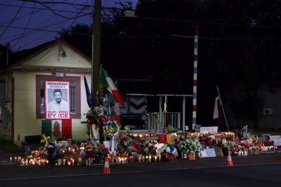 A makeshift memorial with dozens of candles and flowers laid in front of a small house at night.
