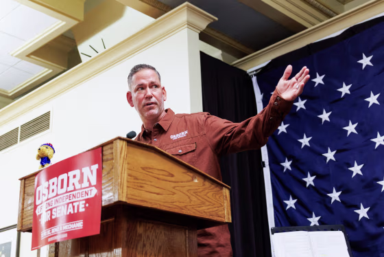 Dan Osborn speaks at a podium and extends his hand outwards, there is a red poster on the front of the podium that reads in white text: "Osborn, lifelong independent, for senate. To fix DC, send a mechanic."