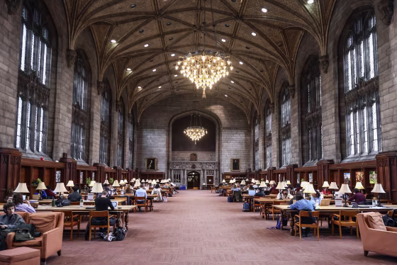 A large stone room with a vaulted ceiling and chandelier and long rows of wooden desks.