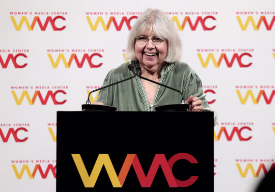 Robin Morgan smiles while standing in front of two microphones on a lectern.
