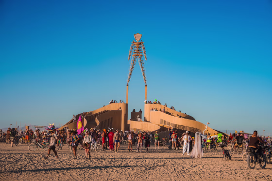 A large wooden man sculpture, The Man, stands in Black Rock Desert, Nevada, surrounded by festival participants in colorful costumes and bicycles