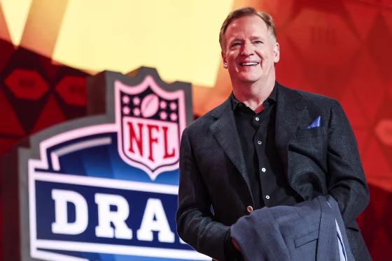 Roger Goodell smiles on stage while holding a folded suit in his hands