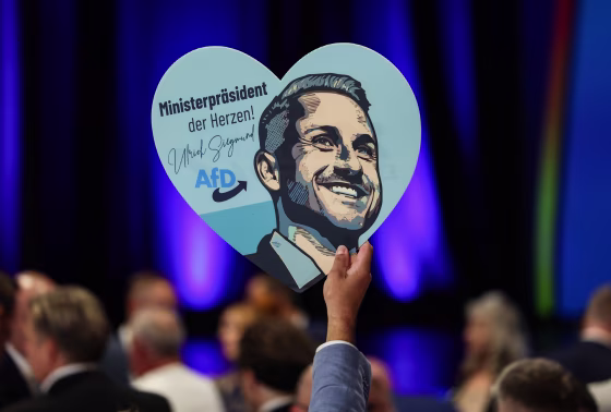 A hand holds up a heart-shaped placard that shows an illustrated depiction of Ulrich Siegmund