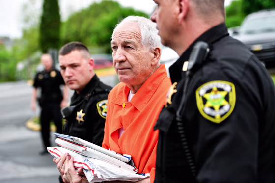 Jerry Sandusky holds paperwork. while walking in between two police officers outside, he wears an orange jumpsuit