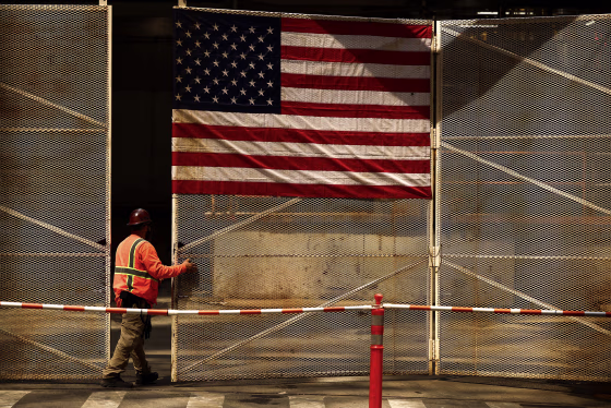 A construction worker wearing an orange vest and hardhat stands beneath a large American flag on a fence.