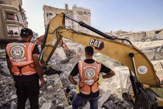 Two men wearing orange vests stand in front of two diggers in a field of debris.