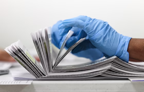 A gloved hand (blue) counts a stack of mail-in ballots on a table, side view