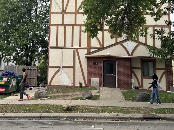 Two pedestrians pass a weathered tan building with a dumpster and litter out front. 