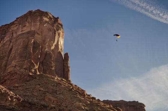 A person floating under a red, black and yellow parachute in front of a large stone cliff.