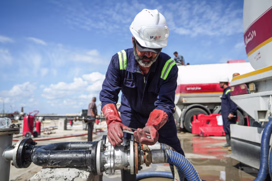 A worker in a hard hat twists the lever of a pipe outside near large trucks getting filled with oil
