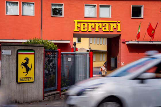 A detail view shows Ferrari factory signage, a yellow shield bearing the black prancing horse and Italian tricolor stripes, mounted on a concrete wall.