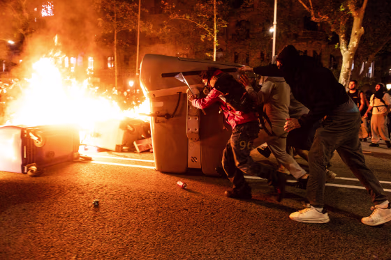 Trash containers burn in a central street at night near protestors